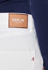 Balti džinsiniai šortai su rudos odos etiketę, ant kurios užrašyta "REPLAY SINCE 1981". Raudonas siuvinėjimas "R" kišenėje.