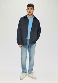 Tmavomodrá bunda s dvoma prednými vreckami, nosená na svetlomodrej košeli, v kombinácii so svetlými džínsy a krémovými teniskami s textúrovanými akcentmi.