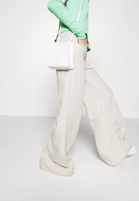 Široké béžové lněné kalhoty ve spojení s živě zeleným dlouhým rukávem. Na rameni visí malá, strukturovaná bílá kabelka.