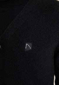 Cardigan noir en tricot avec une petite patte noire ornée d'un logo blanc abstrait sur la poitrine gauche.