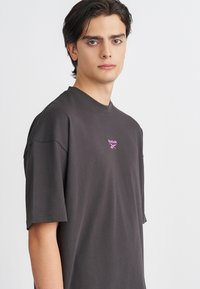 Camiseta de manga corta gris oscuro hecha de algodón suave. Presenta un pequeño logo de Reebok en color rosa en el pecho. Diseño simple y de corte relajado.