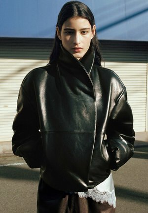 Osoba o długich ciemnych włosach ubrana w czarną, oversize'ową skórzaną kurtkę i białą odzież z koronkową lamówką, stojąca na zewnątrz na tle pasiastej ściany.