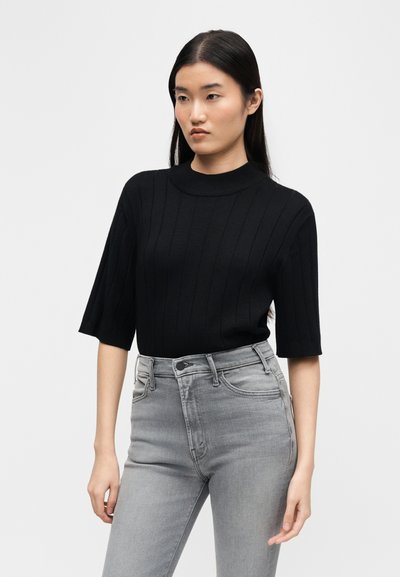 Νεαρή γυναίκα με μακριά σκούρα μαλλιά, που φοράει μαύρο ριμπ κοντομάνικο τοπ και ψηλόμεσα ανοιχτόγκρι τζιν, μπροστά σε απλό φόντο.