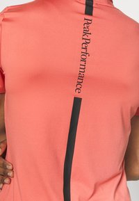 Maglietta da performance corallo con testo verticale nero, maniche corte e superficie liscia. Presenta il logo "Peak Performance" lungo la schiena.