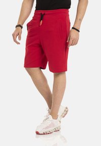 Rode katoenen shorts met een tailleband met verstelkoord, twee zijzakken en een middelmatige lengte. Gedragen met witte en rode sportschoenen.