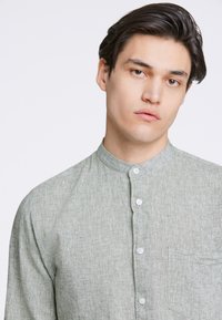 Camicetta button-up verde chiaro, con texture, colletto a stand, taschino frontale e bottoni bianchi, progettata con una vestibilità casual e rilassata.