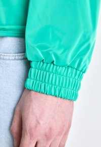 Jacheta de vânt teal cu manșete elasticizate și textură netedă, lucioasă. Fabrică de denim vizibil sub jachetă, arătând o culoare albastru deschis.