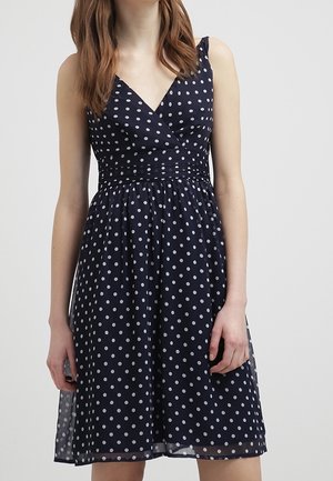 Kobieta nosząca granatową sukienkę bez rękawów w białe kropki z dopasowaną talią, na jednolitym jasnym tle.