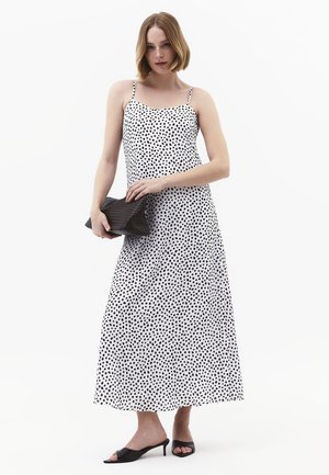 Frau trägt weißes ärmelloses Maxikleid mit schwarzen Punkten, hält eine schwarze strukturierte Clutch und schwarze offene Sandalen mit Absatz.