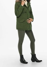 Parka vert olive avec une capuche bordée de fourrure, des poches frontales et une taille à cordon de serrage. Associée à des leggings olives ajustés et des bottines noires jusqu'aux chevilles.