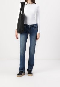 Jean en denim bleu avec une coupe droite, associé à un haut blanc à manches longues ajusté et un sac noir. Les chaussures sont des baskets noires et blanches.