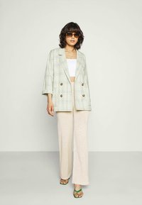 Blazer duplo em padrão xadrez nas cores verde claro e branco, com botões castanhos, combinado com calças largas bejes e sandálias verdes.
