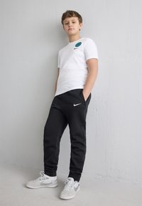 Chlapec v bielom tričku Nike, čiernych teplákoch Nike a bielych teniskách Nike, stojaci sebavedome oproti jednoliatej svetlosivej stene.