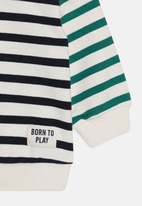Ribadega sweatshirt tumesiniste, valgete ja roheliste triipudega; omab pehmet tekstuuri, soonikuid varrukaotsi ja "BORN TO PLAY" silt.