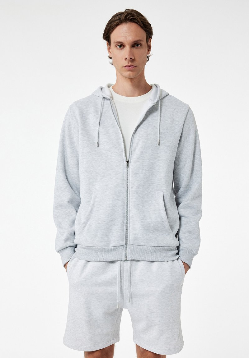 Lichtgrijze rits hoodie gemaakt van zachte stof met een capuchon en trekkoorden. Gepaard met bijpassende shorts met een elastische tailleband.