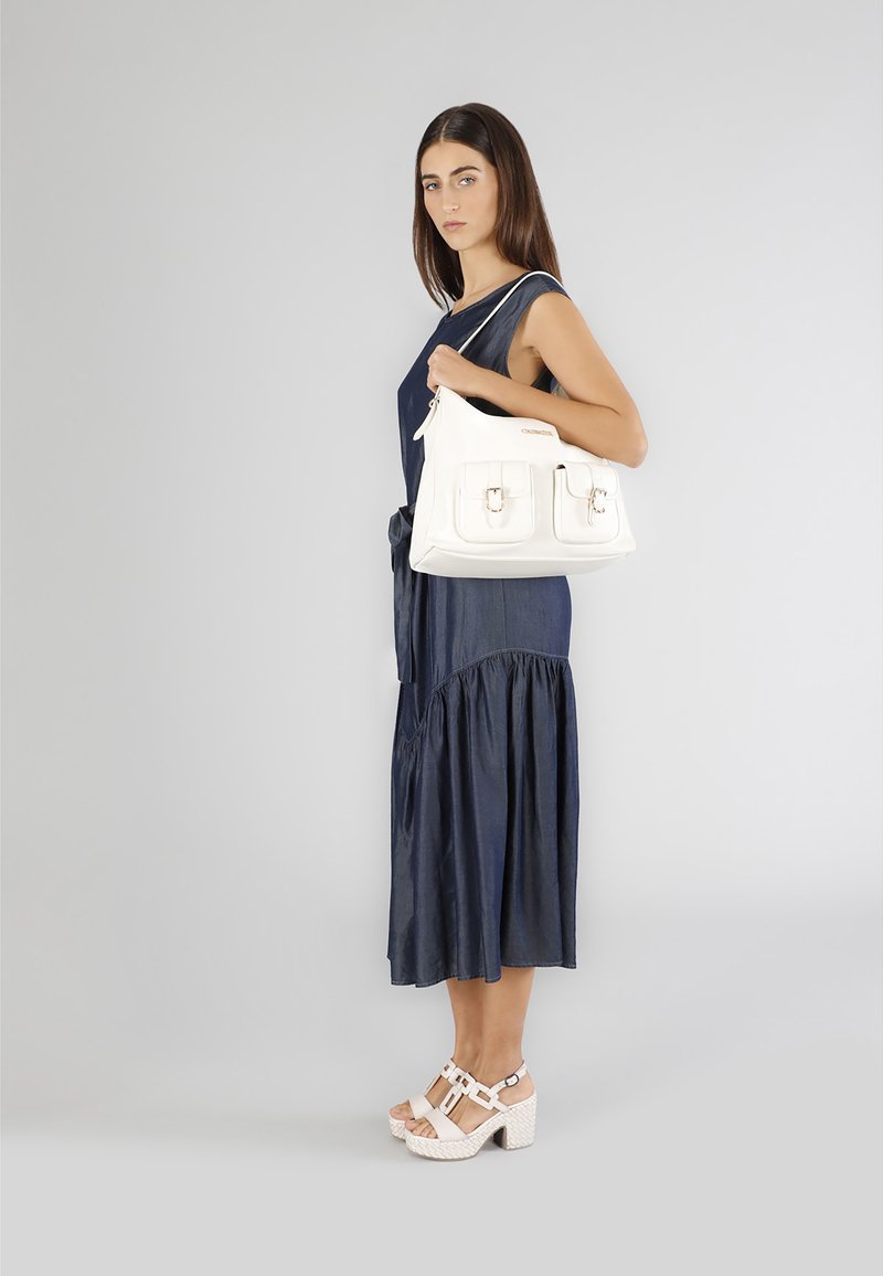 Bolso de mano blanco con dos bolsillos frontales y cierre de cremallera, sostenido por una persona vestida con un vestido sin mangas fluido en color marino y tacones gruesos de color claro.