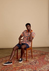 Korte mouwen, koraalkleurige polo met groene en blauwe bloemenpatronen, gecombineerd met marineblauwe shorts en marineblauwe sneakers, zittend op een houten stoel.