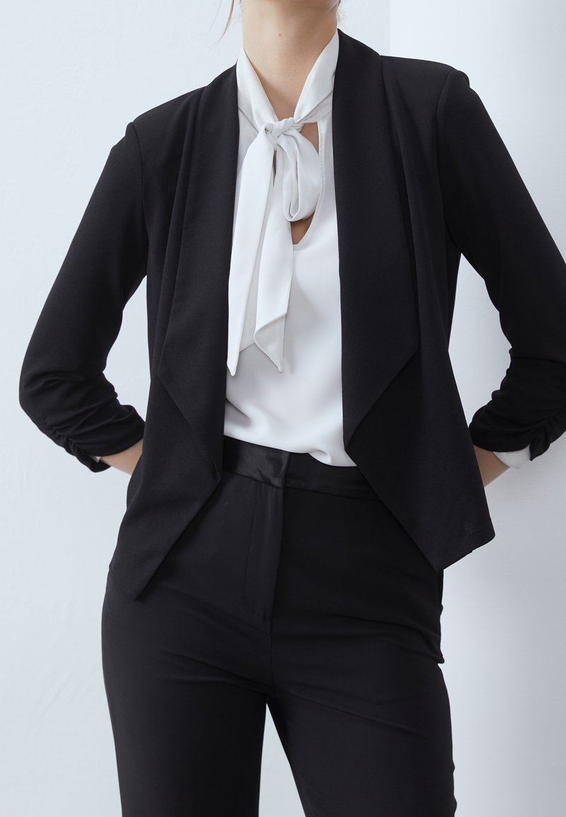 Femme portant un blazer noir cintré, un pantalon noir et une blouse blanche avec un nœud noué au col, debout les mains sur les hanches.