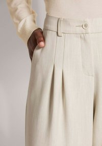 Plissébroek in een lichtbeige stof met een gladde textuur. De tailleband heeft een knoop en zijzakken voor extra gemak.