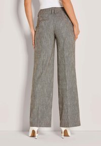 Vrouw in een grijs hoge taille wijde broek en witte hoge hakken, staand tegen een effen lichte achtergrond.