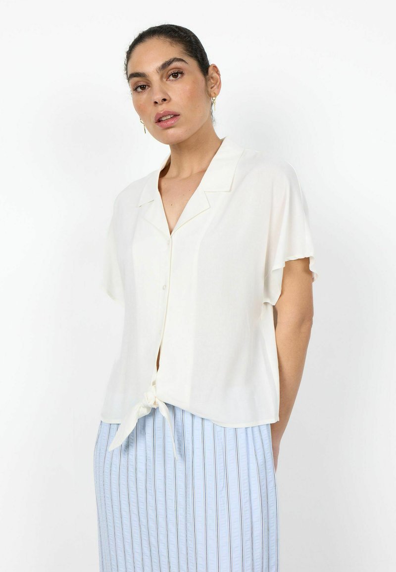 Femme portant un chemisier blanc à manches courtes boutonné, noué à la taille, avec une jupe à rayures verticales bleu clair sur fond blanc.