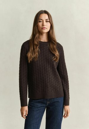 Junge Frau mit langen braunen Haaren, die einen dunkelbraunen Zopfstrickpullover und blaue Jeans trägt und vor einem schlichten hellen Hintergrund steht.