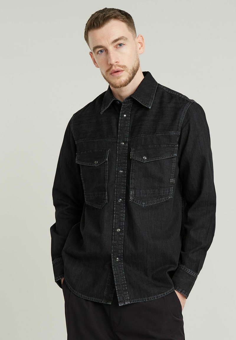 Camisa de mezclilla negra con mangas largas, cierre frontal de botones, dos bolsillos en el pecho y detalles de costura en contraste. Textura suave y corte casual.