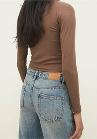 Kobieta w brązowej bluzce z długim rękawem i jasnoniebieskich dżinsach z wysokim stanem, pokazana od tyłu z długimi ciemnymi włosami.