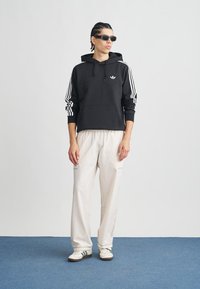 Juodas džemperis su baltais dryžiais ant rankovių ir logotipu, derinamas su šviesios spalvos, plačiomis kelnėmis ir baltais sportbačiais. Dėvimi tamsūs akiniai nuo saulės.