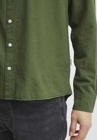 Camisa verde de botón con un tejido texturizado, con botones blancos y una etiqueta lateral, combinada con pantalones gris oscuro. Primer plano de la manga y el dobladillo.