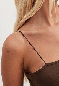 Ruskea, vartalonmyötäinen toppi ohuilla olkaimilla, jossa on jäsennelty kaula-aukko ja sileä pinta, korostaen olkapäitä ja solisluun aluetta.