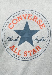 Λογότυπο Converse All Star σε πορτοκαλί και σκούρο μπλε με ένα μεγάλο αστέρι και το κείμενο «Chuck Taylor» σε γκρι υφασμάτινο φόντο.