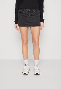 Osoba v černé džínové mini sukni, černém dlouhém rukávu s bílými logy na rukávech, bílých ponožkách Adidas a bílých teniskách.