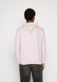 Persona che indossa un hoodie rosa chiaro e jeans grigio scuro, in piedi di fronte a uno sfondo bianco semplice con le spalle rivolte verso la fotocamera.