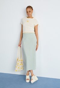 Top court crème avec volants jaunes et détail floral, jupe midi vichy bleu et vert, chaussures compensées crème et sac tote en fleurs tissé.