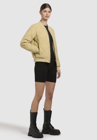 Gequilted beige bomberjack met ribbelboorden en -zoom, gecombineerd met zwarte aansluitende shorts en stevige zwarte veterschoenen.
