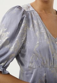 Blusa lavanda con maniche a sbuffo voluminose, caratterizzata da un motivo metallico oro, scollatura a V e dettaglio con bottone davanti. Texture setosa.