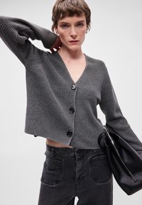 Cardigan cinza canelado com decote em V, apresentando quatro botões pretos e mangas longas, combinado com jeans escuros de cintura alta e uma bolsa preta.