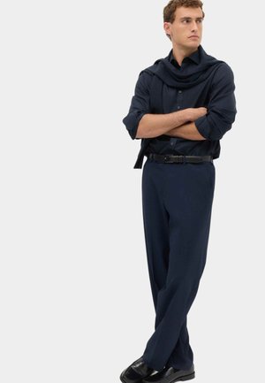 Mand i mørk marineblå skjorte og bukser med en sweater draperet over skuldrene står med armene over kors og ser til siden mod en ensfarvet baggrund.