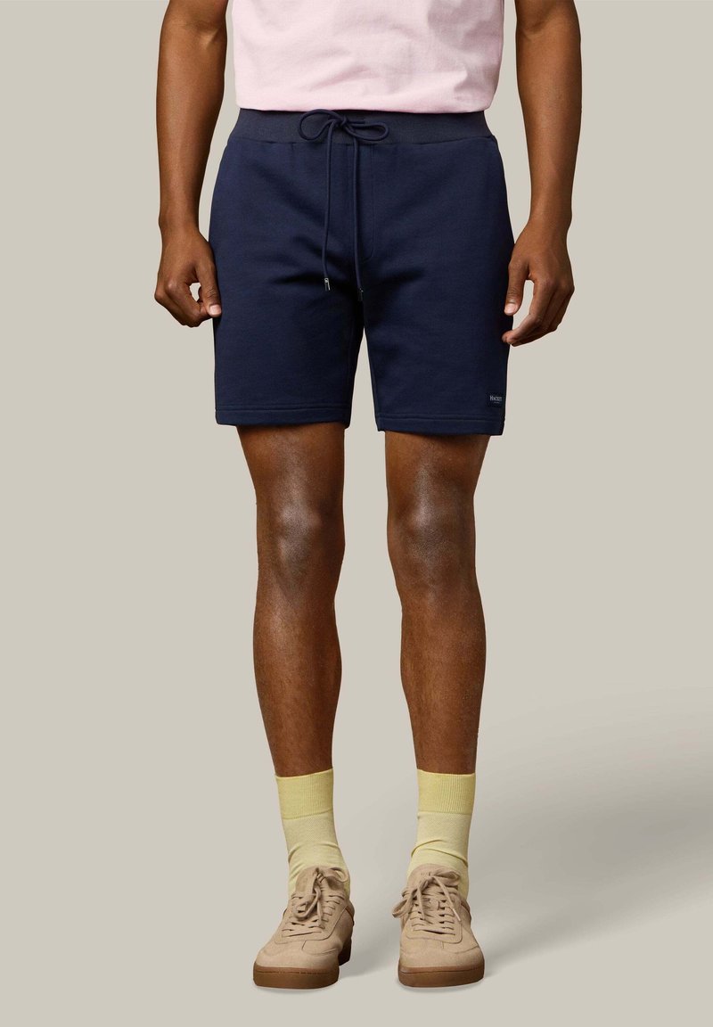 Mężczyzna ubrany w granatowe szorty z ściągaczem, jasnoniebieskie skarpetki, beżowe trampki oraz jasnoróżową koszulę na neutralnym tle.