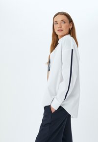 Witte longsleeve shirt met een ontspannen pasvorm, voorzien van marineblauwe streepaccenten op de mouwen en een knoopsluiting bij de hals. Gaat gepaard met marineblauwe broeken.