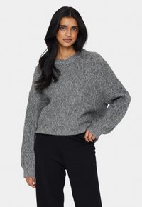Gestrickter Pullover in hellgrau mit einem Kabelmuster, Rundhalsausschnitt und lockerer Passform. Rippbündchen und Saum sorgen für Textur. Kombiniert mit schwarzen Hosen.