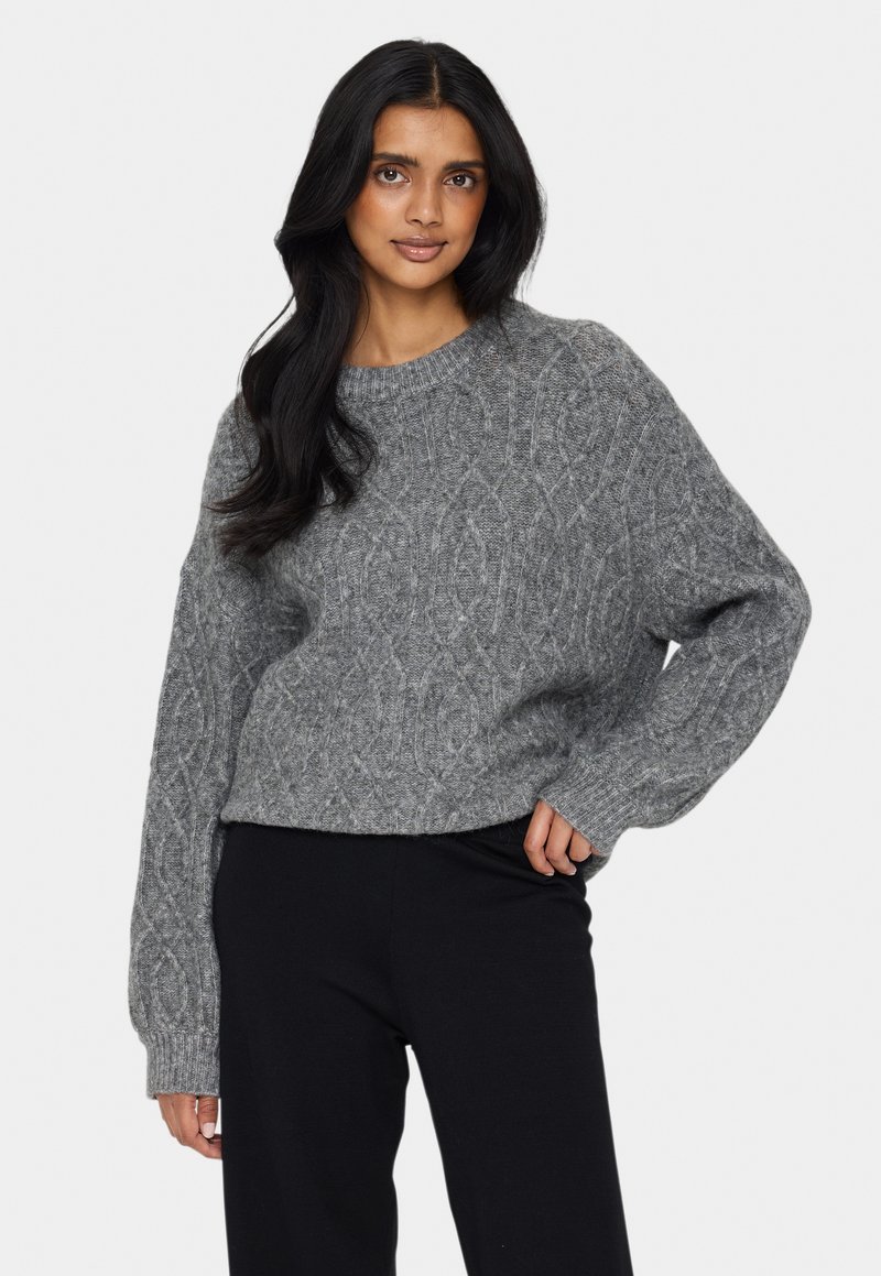 Gestrickter Pullover in hellgrau mit einem Kabelmuster, Rundhalsausschnitt und lockerer Passform. Rippbündchen und Saum sorgen für Textur. Kombiniert mit schwarzen Hosen.