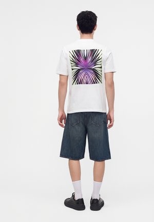 Man draagt een wit t-shirt met paars en zwart abstract ontwerp op de rug, donkere knielange spijkershorts, witte sokken en zwarte schoenen.