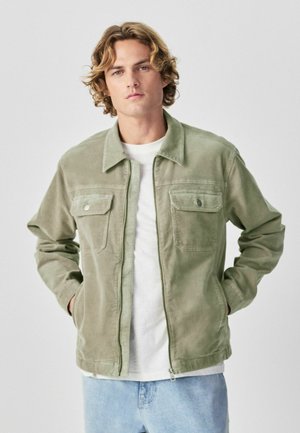 Junger Mann mit lockigem blondem Haar, der eine hellgrüne Cordjacke, ein weißes T-Shirt und hellblaue Jeans vor einem einfachen Hintergrund trägt.