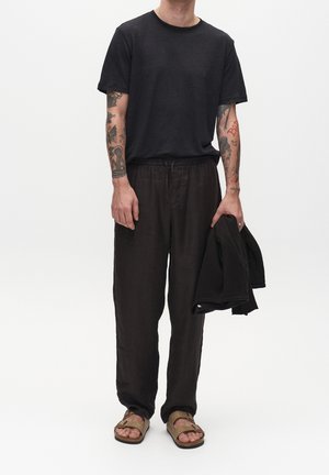 T-shirt nero a maniche corte, pantaloni larghi in lino marrone, sandali marroni, che tiene una giacca nera, sfondo neutro.