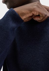 Jersey de lana azul marino con un acabado texturizado y esponjoso. Primer plano de una mano descansando sobre el hombro, destacando los detalles del tejido y el diseño de los puños.