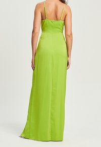 Kobieta stojąca tyłem, ubrana w zieloną, satynową suknię do kostek z cienkimi, regulowanymi ramiączkami.