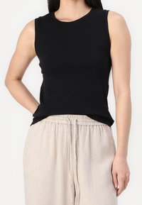 Mujer vestida con un top negro sin mangas metido dentro de pantalones beige con cordón, de pie con una mano en el bolsillo contra un fondo blanco.