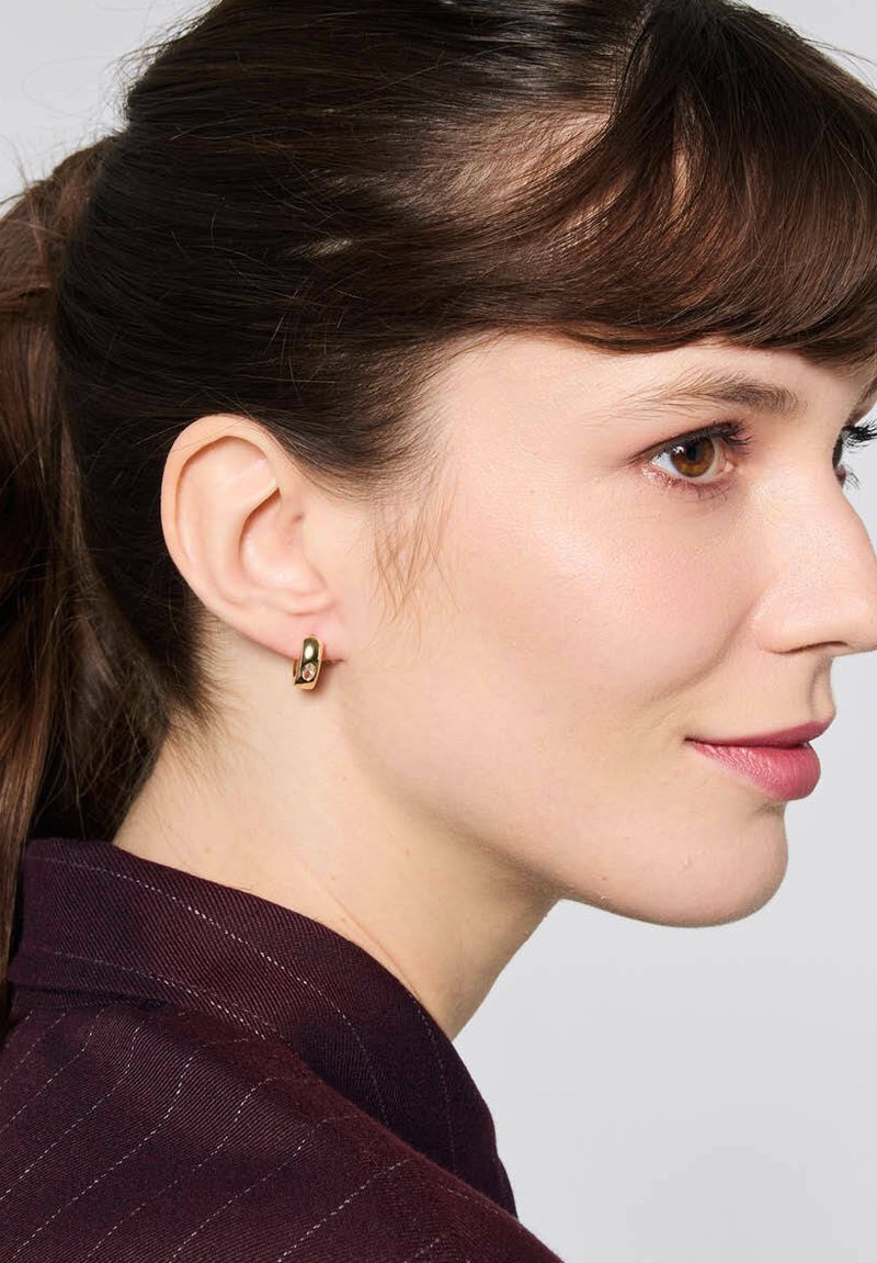 Złote kolczyki w kształcie kół, z gładkim wykończeniem i małym akcentującym kamieniem. Noszone na lewym uchu, pokazane obok ciemnoczerwonej bluzki.